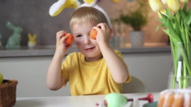 Emotional portrait of a cheerful little boy wearing bunny ears on Easter day who laughs merrily, plays with colorful Easter eggs sitting at a table in the kitchen.