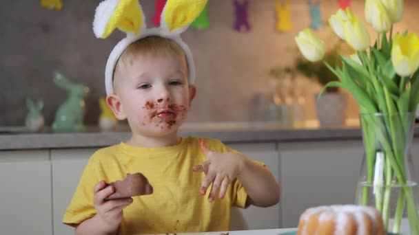 Маленький Милый Мальчик Кроличьими Ушами Ест Шоколадного Пасхального Кролика Сидя — стоковое видео