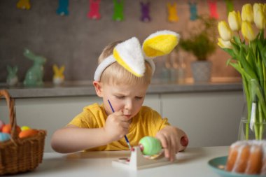 A little boy wearing bunny ears prepares painted Easter eggs for Easter while sitting at the kitchen table. Happy easter.