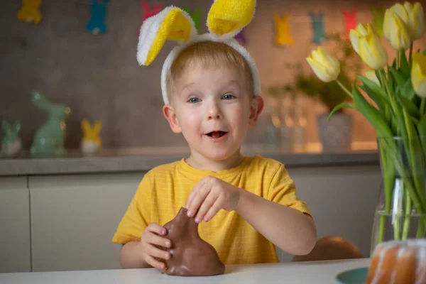 A cute little boy wearing bunny ears on Easter day is eating a chocolate Easter bunny. A child plays egg hunt for Easter. Charming child celebrates Easter at home.