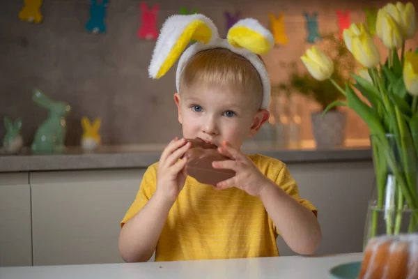 A cute little boy wearing bunny ears on Easter day is eating a chocolate Easter bunny. A child plays egg hunt for Easter. Charming child celebrates Easter at home.