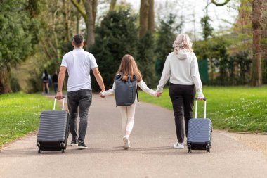 Üç kişilik bir aile, baba, anne ve kız, bavullarla parkta yürüyorlar..