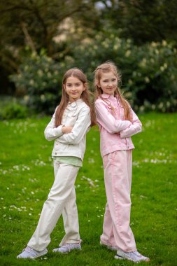 Açık renkli elbiseli, gülümseyen, yeşil çimlerin üzerinde duran iki arkadaş canlısı kız parkta bahar gününün tadını çıkarıyor.