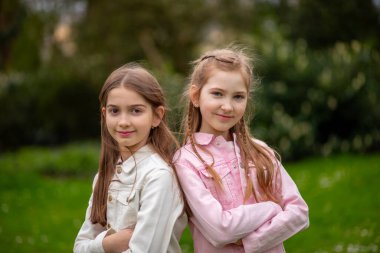 Pastel gölgeli giysiler içinde iki büyüleyici kız, gülümsüyor ve yeşil bir bahar parkında poz veriyorlar.