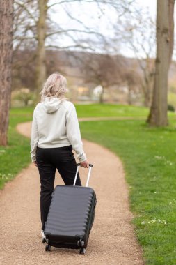 Park yolunda bavulu taşıyan bir kadın, kameradan yüzünü çevirmiş, yandan görülmüş.