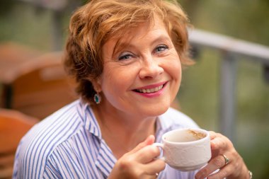 Neşeli bir kadın elinde kahve fincanıyla balkonda içkisini yudumlarken yüzünü aydınlatan yumuşak güneş ışığının tadını çıkarıyor..