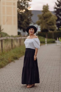 Tarz sahibi bir kadın kaldırım taşından bir patikada duruyor, omuzları açık beyaz bir elbise ve dalgalı siyah bir etek içinde, zarif bir şekilde parıldıyor..