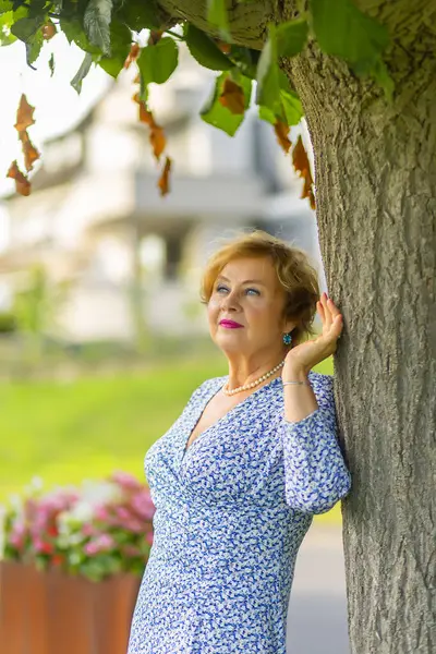 stock image A woman stands by a tree, enjoying the sunshine, wearing a blue patterned dress and accessorized with elegant earrings.