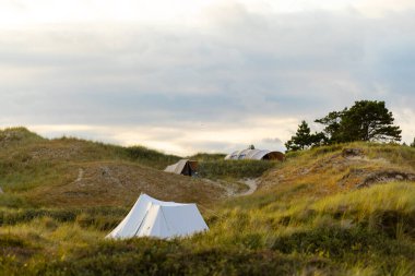 A peaceful campsite is set up among grassy dunes as the sun begins to set. Tents are positioned on a winding trail, surrounded by lush vegetation and a serene sky. clipart