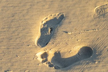 Sıcak kuma iki ayak izi kazınır, batan güneşin altında yumuşak bir şekilde parlar, sahilde huzurlu bir atmosferi yansıtır..