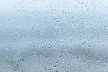 Yağmur damlaları bir pencerede toplanır ve ötesindeki manzarayı bozar. Hava bulutlu, içeriden yumuşak, sessiz bir ışık yayılıyor ve huzurlu bir ortam yaratıyor..