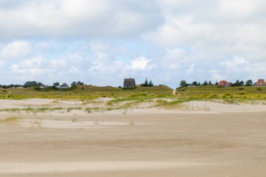 Bir manzara uzak evlere giden kumlu kumsalları gösteriyor. Sahne bulutlu bir gökyüzünün altında, kıyı bölgesinin sükunetini vurguluyor..