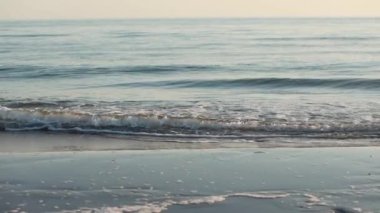 Yumuşak okyanus dalgaları sürekli kıyıya vurur ve yaz sonu sıcak gökyüzünde güneş batarken kumlu bir plajda huzurlu bir atmosfer yaratır..