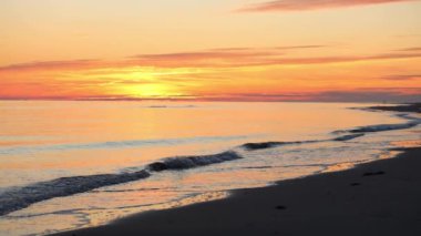 Sakin bir günbatımı gökyüzünü canlı renklerle boyarken yumuşak dalgalar kumlu sahilde süzülüyor ve rahatlamak için mükemmel sakin bir atmosfer yaratıyor..