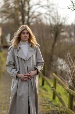 Genç bir kadın ağaçlar ve suyla kaplı huzurlu bir patikada yürüyor. Şık bir trençkot giyiyor ve doğa atmosferinin tadını çıkarırken yansıtıcı görünüyor..