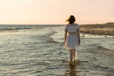 Çiçekli elbiseli bir kadın gün batımında sığ sularda gezer. Yumuşak dalgalar, güneş ufkun altına batarken sakin bir atmosfer yaratıyor..
