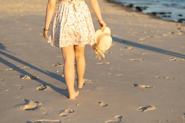 Çiçekli elbiseli bir kadın gün batımında kumlu bir plajda yürür, hasır bir şapka taşır ve kumsalda ayak izleri yaratır,.