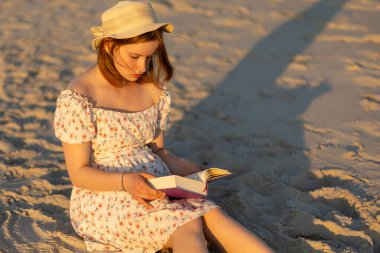 Kumlu sahilde oturan, çiçekli bir elbise ve hasır şapka giyen genç bir kadın kendini bir kitaba daldırıyor. Güneş batıyor, etrafında sıcak altın bir ışık saçıyor..