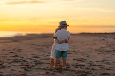 Çocuklar kumlu sahilde birbirlerine yakın dururlar. Birbirlerine sarılırlar. Güzel bir günbatımı gökyüzü ve okyanus boyunca ılık renkler bırakır..