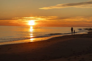 Güneş sakin okyanusun üzerinde batarken kumlu bir sahil boyunca el ele dolaşırlar. Gökyüzünün canlı renkleri suyu yansıtıyor ve huzurlu bir atmosfer yaratıyor..