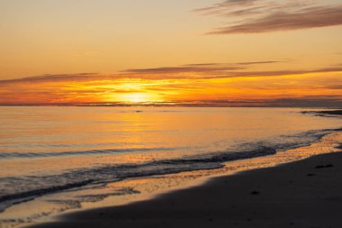 Nefes kesen bir günbatımı, turuncu ve pembe parlak renkler ile gökyüzünü kumlu bir kıyıya doğru hafif dalgalar gibi boyar. Huzurlu sahne rahatlamayı ve yansımayı davet eder..