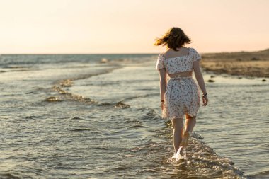 Genç bir kadın gün batımında sahildeki sığ sularda yalın ayak dolaşır. O sakin denize ve ufka hayranlık duyarken elbisesi hafif meltemde dalgalanıyor..