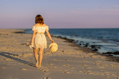 Gün batımında, elinde hasır bir şapkayla çiçekli bir yaz elbisesi giymiş bir kadın kumsalda yalınayak dolaşıyor. Sakin deniz gökyüzünün sıcak renklerini yansıtır..