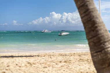 İki tekne turkuaz sularda sessizce yüzer, ön planda kumlu bir plaj ve yukarıdaki parlak mavi gökyüzüne yayılmış tüylü bulutlarla. Huzurlu bir sahil atmosferi hakim..
