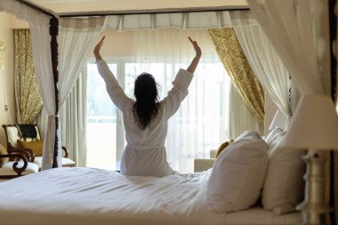 Bornoz giyen bir kadın doğal ışıkla dolu lüks bir otel odasında kollarını açıyor. Güneş ışığı perdelerin arasından süzülür, sakin bir sabah atmosferi yaratır..