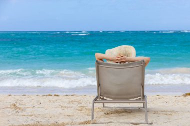 Güneş şapkalı bir insan plajdaki bir sandalyede rahatça uzanır, dalgaların ve sıcak güneşin rahatlatıcı sesinin tadını çıkarır. Dalgalı okyanus uzaklarda uzanıyor..