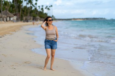 Bir kadın kumlu bir plajda yürüyor, ayakları ılık havanın tadını çıkarırken yumuşak dalgalarda. Tomurcuklu palmiye ağaçları ve yumuşak dalgalar etrafında sakin bir atmosfer yaratıyor..