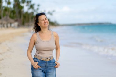 Gülümseyen bir kadın güzel bir sahilde yürüyor, ayaklarının altındaki yumuşak kumu hissediyor. Gökyüzü açık ve sakin dalgalar, güneşli bir öğleden sonra kıyıda yavaşça süzülüyor..