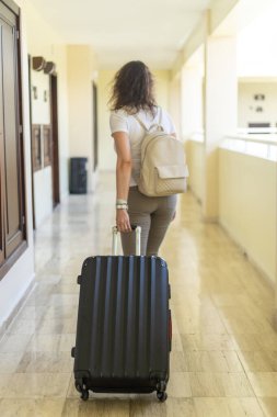 Bir kadın otel koridorunda yürürken görülüyor, bavulunu sırtında taşıyor ve bir sırt çantası taşıyor. Parlak salonda modern tasarım elementleri ve doğal ışık yer alıyor..