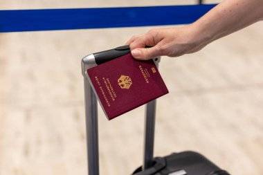 Havaalanındaki bir bavul sapında dinlenirken bir el pasaport tutuyor. Mavi kontrol engeli gezginleri, yoğun bir seyahat gününü gösterir.