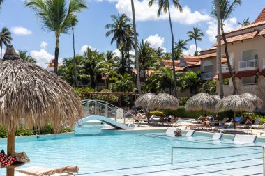 Punta Cana, Dominik Cumhuriyeti - Mart 24, 2025: Konuklar güneşli bir öğleden sonrayı tropikal tatil beldelerinde güneşli bir havuzda, sazdan şemsiyeler altında uzanarak, palmiye ağaçları ve canlı yeşilliklerle çevrili.