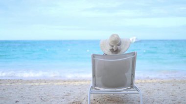 Plajda huzurlu bir günün tadını çıkaran bir insan, geniş kenarlı bir şapka takmış bir sandalyede oturuyor. Işık dalgaları, parlak bir gökyüzünün altında, sakin bir atmosfer sağlar..