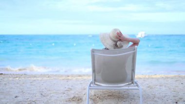 Bir kadın plaj sandalyesinde, geniş kenarlı bir şapka takarak, güneşlenerek ve ferahlatıcı okyanus esintisiyle huzur içinde oturur. O dinlenirken dalgalar yavaşça arka planda çarpar.