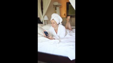 Kafasına havlu sarılmış bornozlu bir kadın otel yatağında dinleniyor. Akıllı telefonuna odaklanmış, sakin bir atmosferde boş zamanlarının tadını çıkarıyor..