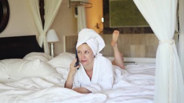 Yatağında yumuşak bir bornoz ve havluyla telefonda konuşan bir kadın var. Davet edilen yatak odası doğal ışıkla dolu. Huzurlu bir ortam yaratıyor..