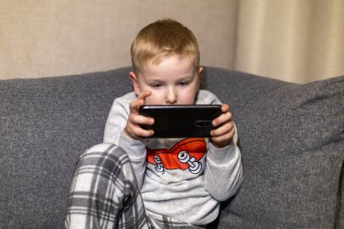 Rahat bir pijama giymiş genç bir çocuk yumuşak bir kanepede otururken dikkatle akıllı bir telefona odaklanıyor. Atmosfer evde rahat bir akşamı yansıtıyor..