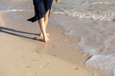 Bir insan kumlu sahilde yürüyor, sıcak güneşin ve ayaklarına vuran nazik dalgaların tadını çıkarıyor. Sahne, okyanus kıyısında sakin bir anı yakalar..