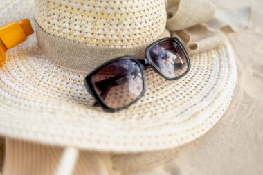 Kurdeleyle süslenmiş geniş kenarlı hasır bir şapka, güneşli bir plaj gezisi için mükemmel, şık siyah güneş gözlüğü ve güneş kreminin yanında sıcak kumun üzerinde duruyor..