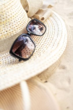 Bir çift siyah güneş gözlüğü yumuşak, altın kum üzerinde geniş kenarlı bir hasır şapkaya gelişigüzel yerleştirilir. Parlak güneş batıyor, rahatlatıcı bir yaz ortamı yaratıyor..