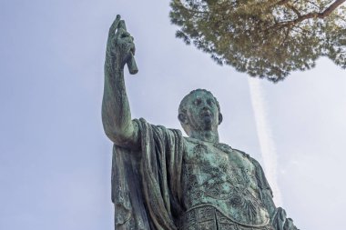 Roma İmparatoru 'nun detaylı bronz heykeli parşömeni yüksek tutuyor ve gök mavisi arkaplana hükmeden bir varlık sergiliyor. Çevreleyen yeşillik tarihi mekana canlılık katıyor.