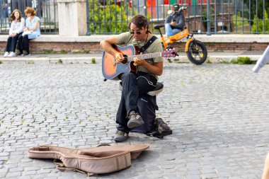 Rom, Italien - 23 Nisan 2025: Bir müzisyen akustik gitarını çalar, yoldan geçenleri büyüler. Canlı, tarihi bir meydanda, yanında bir gitar kılıfı ve güneşin altında rahat bir şekilde oturuyor..