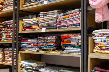 Raflar çeşitli renk ve tasarımlarla düzgünce katlanmış kumaşlarla doludur. Yumuşak ışık desenleri ve desenleri vurgular, müşterileri seçeneklerini keşfetmeye davet eder.