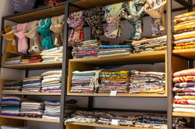 Çeşitli kumaşlar canlı bir dükkanda ahşap raflarda düzenli olarak düzenlenir. Düzenlemede, tasarım ve dikiş projeleri için mükemmel renkli malzemeler sergileniyor.