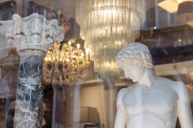 Klasik bir heykel, gün batımının yumuşak tonlarını yansıtan bir dükkan vitrininde duruyor. Arkasında, ışıl ışıl bir avize ve dekoratif bir sütun şehir düzenine cazibe katıyor..