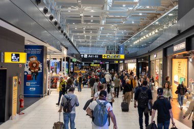 Fiumicino, Rom, Italien - 24 Nisan 2025: Gezginler kalabalık bir havaalanı terminalinde, bagajlarını taşıyarak ve kapılarına doğru ilerliyorlar. Dükkanlar yürüyüş yoluna dizilir, öğleden sonranın hareketli atmosferini artırır..