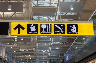 Fiumicino, Rom, Italien - 24 Nisan, 2025: Bir havaalanı terminalinde açık sarı bir tabela alışveriş, yemek seçenekleri, sigara içme alanı ve havayolu yolcu konforu için rehberlik eden bir havayolu salonu simgeleri sergiliyor.
